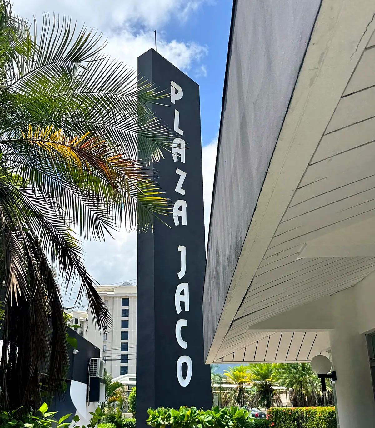 Tótem de Plaza Jacó visto desde abajo, con el nombre en vertical entre palmeras, en Jacó Norte.